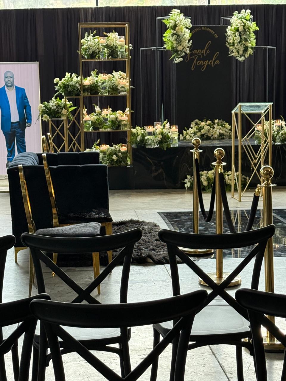 Memorial hall with black cross-back chairs, gold stanchions and floral tribute stage