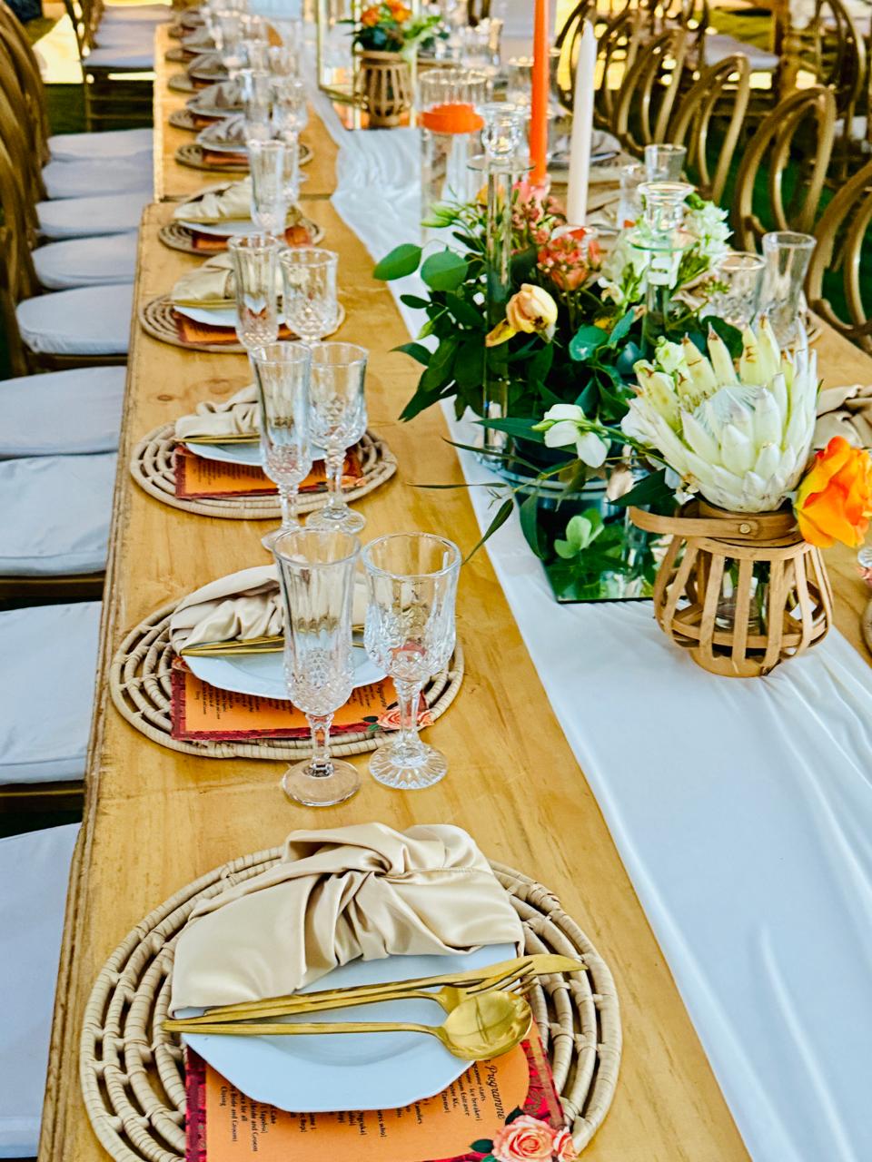 Long wooden banquet table with rattan placemats, gold cutlery and protea centrepieces